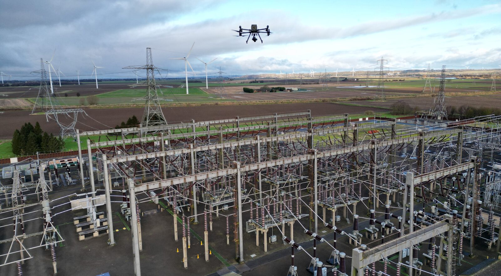 Enterprise drone inspecting a high-voltage substation with wind turbines behind